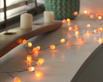 May include: A string of warm white fairy lights with small, round, peach-colored bulbs. The lights are strung on a thin wire and are draped over a white surface.
