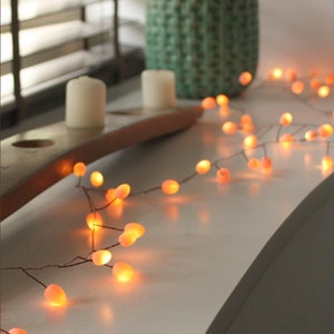 Könnte beinhalten: Eine Lichterkette mit warmweißen Lichtern und kleinen, runden, pfirsichfarbenen Glühbirnen. Die Lichter sind auf einem dünnen Draht aufgefädelt und über eine weiße Oberfläche gelegt.