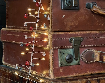 May include: A string of fairy lights with clear and red glass bulbs wrapped around three vintage brown leather suitcases.
