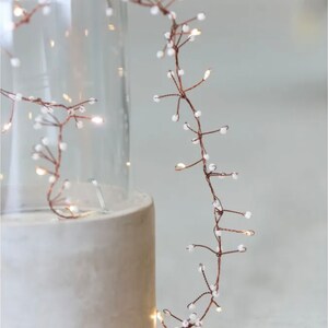 May include: A string of copper wire fairy lights with small white beads. The lights are wrapped around a clear glass vase and a concrete cylinder.