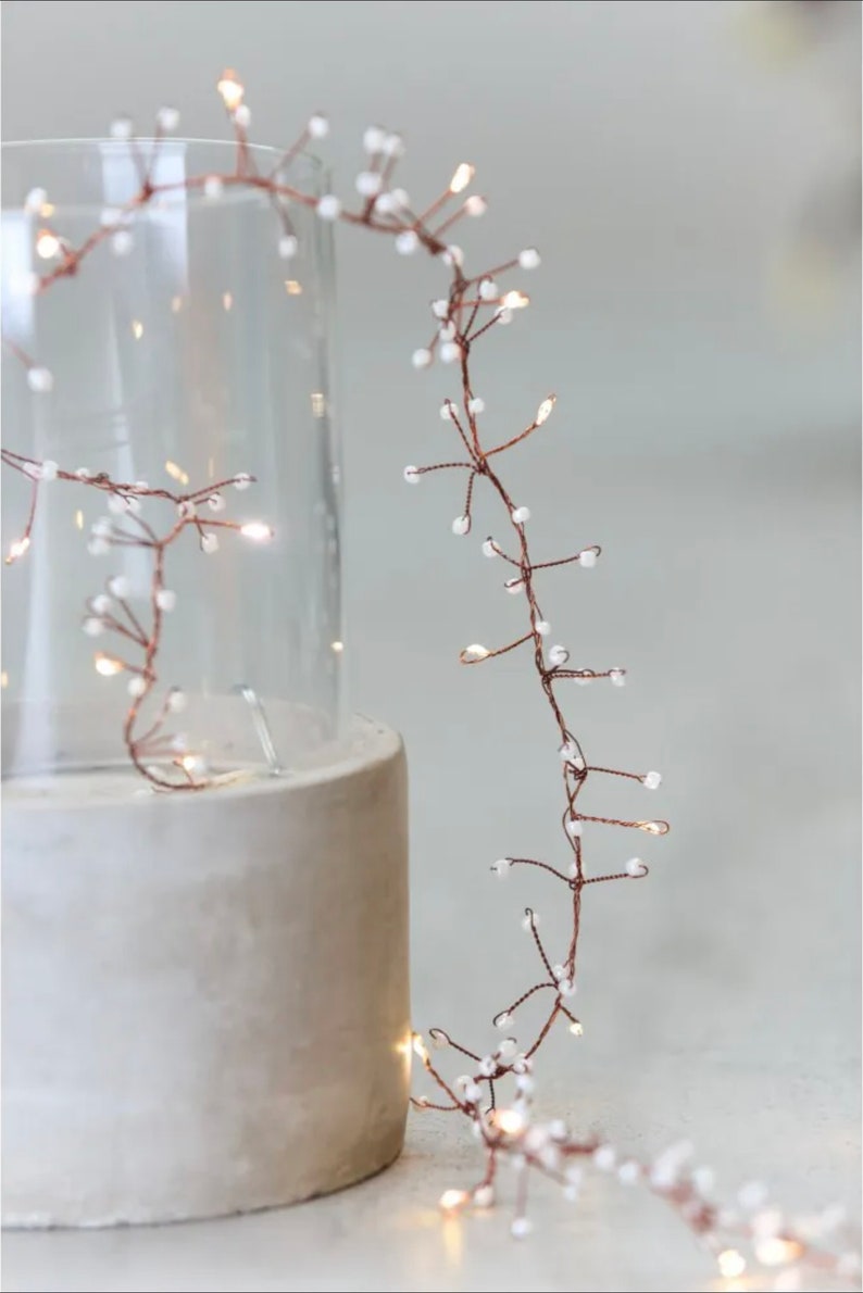 May include: A string of copper wire fairy lights with small white beads. The lights are wrapped around a clear glass vase and a concrete cylinder.