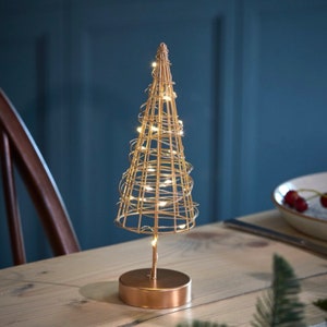 Könnte beinhalten: Ein goldfarbener Weihnachtsbaum aus Draht mit warmweißen LED-Lichtern. Der Baum steht auf einem runden goldenen Sockel und befindet sich auf einem Holztisch.