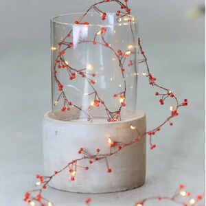 May include: A string of fairy lights with small red beads wrapped around a clear glass cylinder on a concrete base. The lights are strung on a copper wire and create a warm glow.