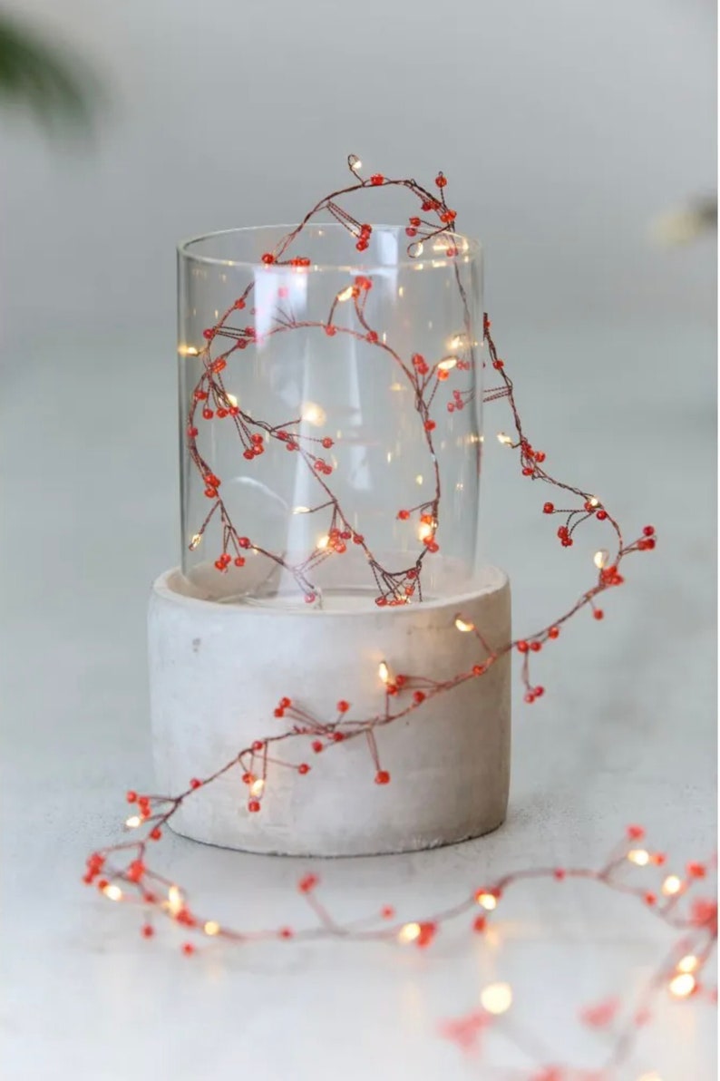 May include: A string of fairy lights with small red beads wrapped around a clear glass cylinder on a concrete base. The lights are strung on a copper wire and create a warm glow.