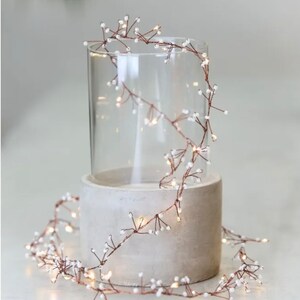 May include: A string of copper wire fairy lights with small white beads. The lights are wrapped around a clear glass cylinder and a concrete cylinder.