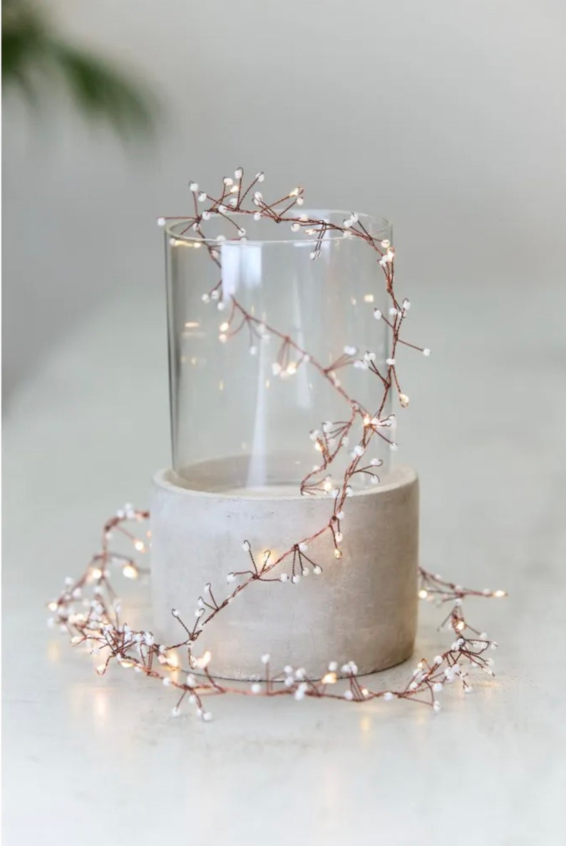 May include: A string of copper wire fairy lights with small white beads. The lights are wrapped around a clear glass cylinder and a concrete cylinder.