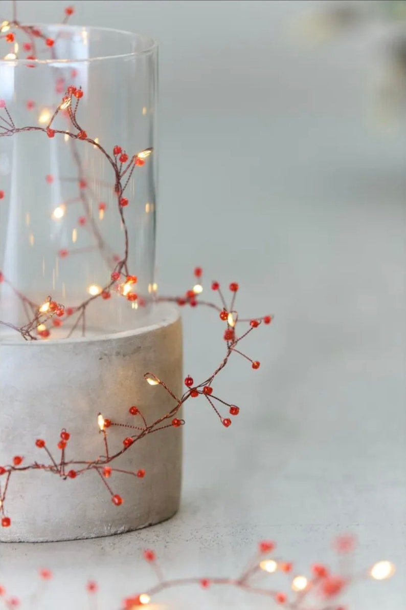 May include: Copper wire fairy lights with small red beads wrapped around a clear glass vase and a concrete cylinder. The lights are strung in a whimsical, cascading pattern.