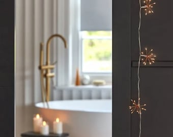 May include: A string of copper fairy lights with starburst shapes, hanging on a dark gray wall. The lights are on and casting a warm glow. The lights are in focus, while the background is blurred.