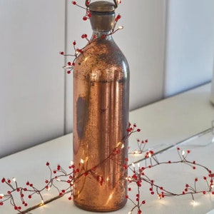 May include: A copper colored bottle with a string of fairy lights wrapped around it. The lights have small red beads along the wire.