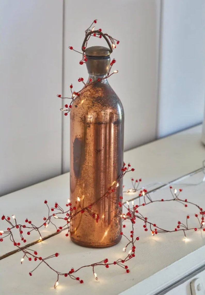 May include: A copper colored bottle with a string of fairy lights wrapped around it. The lights have small red beads along the wire.