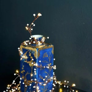 May include: A string of copper wire fairy lights with white beads wrapped around a blue and gold ceramic vase. The lights are lit and casting a warm glow.
