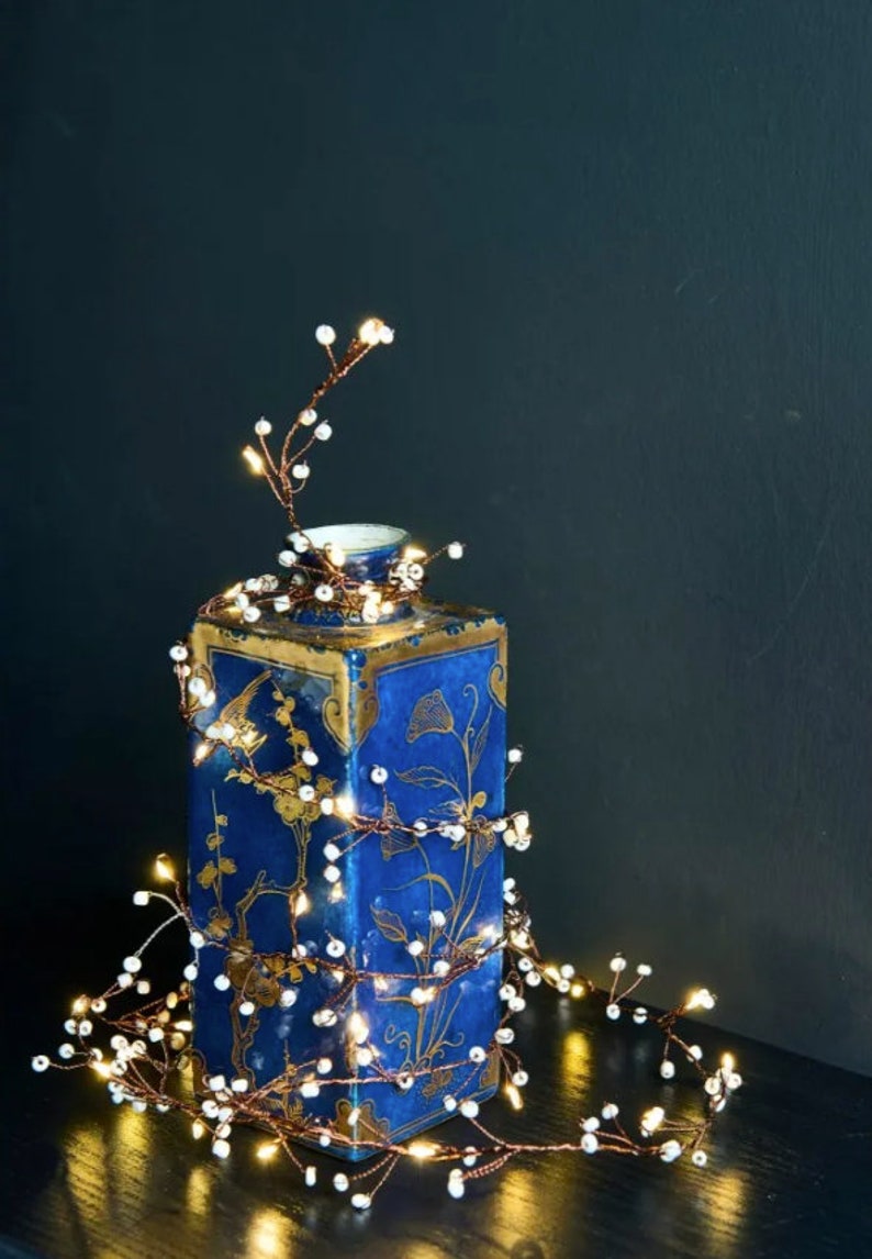 May include: A string of copper wire fairy lights with white beads wrapped around a blue and gold ceramic vase. The lights are lit and casting a warm glow.
