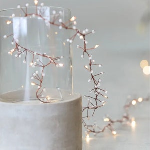 May include: A string of copper wire fairy lights with small white beads, wrapped around a clear glass cylinder on a concrete base.