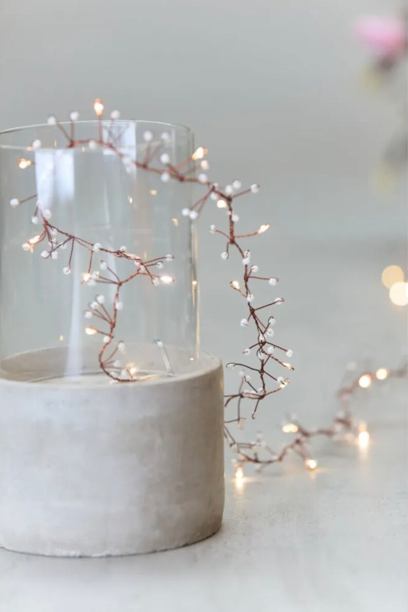 May include: A string of copper wire fairy lights with small white beads, wrapped around a clear glass cylinder on a concrete base.
