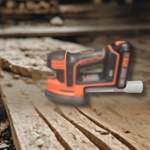 May include: A black and orange cordless sander with a white attachment. The sander is resting on a wooden surface. The tool has a rounded shape and is designed for sanding wood or other materials.