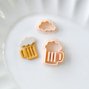 Puede incluir: Tres cortadores de galletas: una nube, una jarra de cerveza y una jarra de cerveza con espuma. Los cortadores son rosados y amarillos.