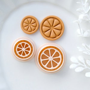 May include: Four orange slice shaped cookie cutters, two are a light brown colour and two are a light peach colour. The cookie cutters are on a white plate.