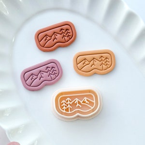 May include: Three cookie cutters in the shape of a pair of ski goggles with a mountain and pine tree design. The cutters are in shades of pink, orange, and white.