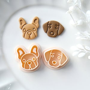 Puede incluir: Cuatro cortadores de galletas de plástico con forma de cara de perro. Dos son marrones y dos son rosas. Los cortadores están en un plato blanco con borde ondulado.