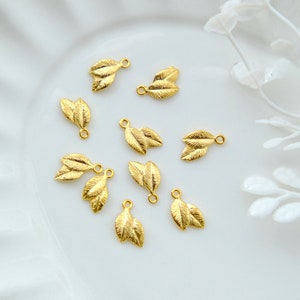 May include: A collection of ten gold leaf charms, each featuring two leaves, arranged on a white plate. The charms have a delicate, intricate design and are perfect for adding a touch of elegance to jewellery or crafts.