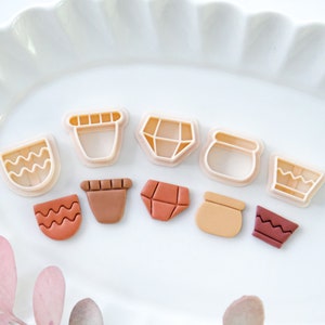 Pode incluir: Um conjunto de dez cortadores de biscoitos em várias formas, incluindo vasos, diamantes e linhas onduladas. Os cortadores são feitos de plástico branco e estão dispostos em um prato branco. Alguns dos cortadores estão cheios de argila marrom.