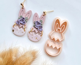 May include: A pair of pink bunny-shaped earrings with purple flowers and gold accents. The earrings are made of polymer clay and are designed to look like a bunny peeking out of an Easter egg. A pink plastic bunny-shaped cookie cutter is also shown in the image.