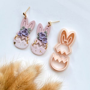 May include: A pair of pink bunny-shaped earrings with purple flowers and gold accents. The earrings are made of polymer clay and are designed to look like a bunny peeking out of an Easter egg. A pink plastic bunny-shaped cookie cutter is also shown in the image.