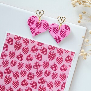 May include: A pair of pink heart-shaped earrings with a gold heart charm. The earrings are decorated with a pink and white strawberry pattern.