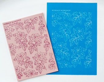 May include: Two sheets of clear plastic with a rose pattern. The sheet on the left is pink with a dark red rose pattern. The sheet on the right is blue with a white rose pattern. Both sheets have the text "Roseaux Workshop" printed on them.