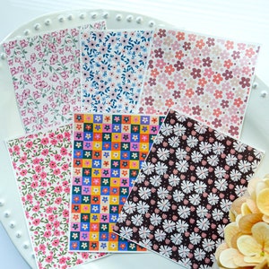 May include: Six different patterned sheets of paper with floral and geometric designs. The patterns include pink flowers on a white background, blue flowers on a white background, pink and red flowers on a white background, colourful squares with flowers on a yellow background, white flowers on a brown background, and pink flowers on a white background.