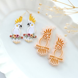 May include: A pair of white cockatoo earrings with yellow crests and floral accents, and two cockatoo cookie cutters in a light peach color.