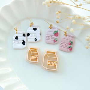 Pode incluir: Quatro pares de brincos de caixa de leite. Dois pares são brancos com manchas de vaca pretas e a palavra "MILK" impressa neles. Um par é rosa com um desenho de morango e a palavra "MILK" impressa neles. O último par é bege com a palavra "MILK" impressa neles.