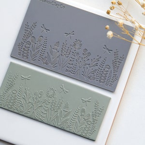 May include: Two rectangular silicone stamps with a floral design and dragonflies. The top stamp is gray and the bottom stamp is green. The text "RoseauxClayCo" is printed on the top stamp.