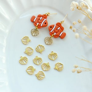 May include: A pair of orange and white clownfish earrings with gold shell charms. The earrings are displayed on a white plate with additional gold shell charms.