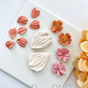 May include: A set of polymer clay flower petals and flowers in shades of pink, orange, and white. The petals are arranged on a white surface with the text "Roseaux Clay Co" in black script.