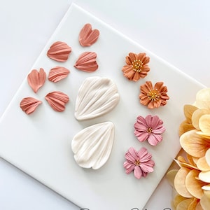 May include: A set of polymer clay flower petals and flowers in shades of pink, orange, and white. The petals are arranged on a white surface with the flowers below. The text "Roseaux Clay Co." is visible in the bottom right corner.
