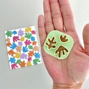 May include: A green plastic stamp with three brown leaf-shaped cutouts. The stamp is held in a person's hand.