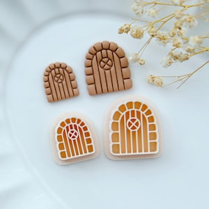 May include: Three brown and orange clay door charms with arched tops and a small window in the center. The charms are different sizes and are arranged on a white plate.