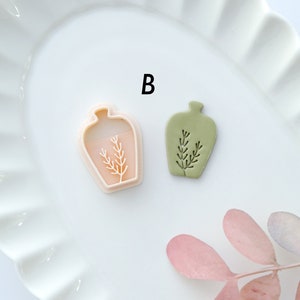 May include: Two ceramic vases with a plant design. The vase on the left is pink and the vase on the right is green. The letter "B" is visible in the background.