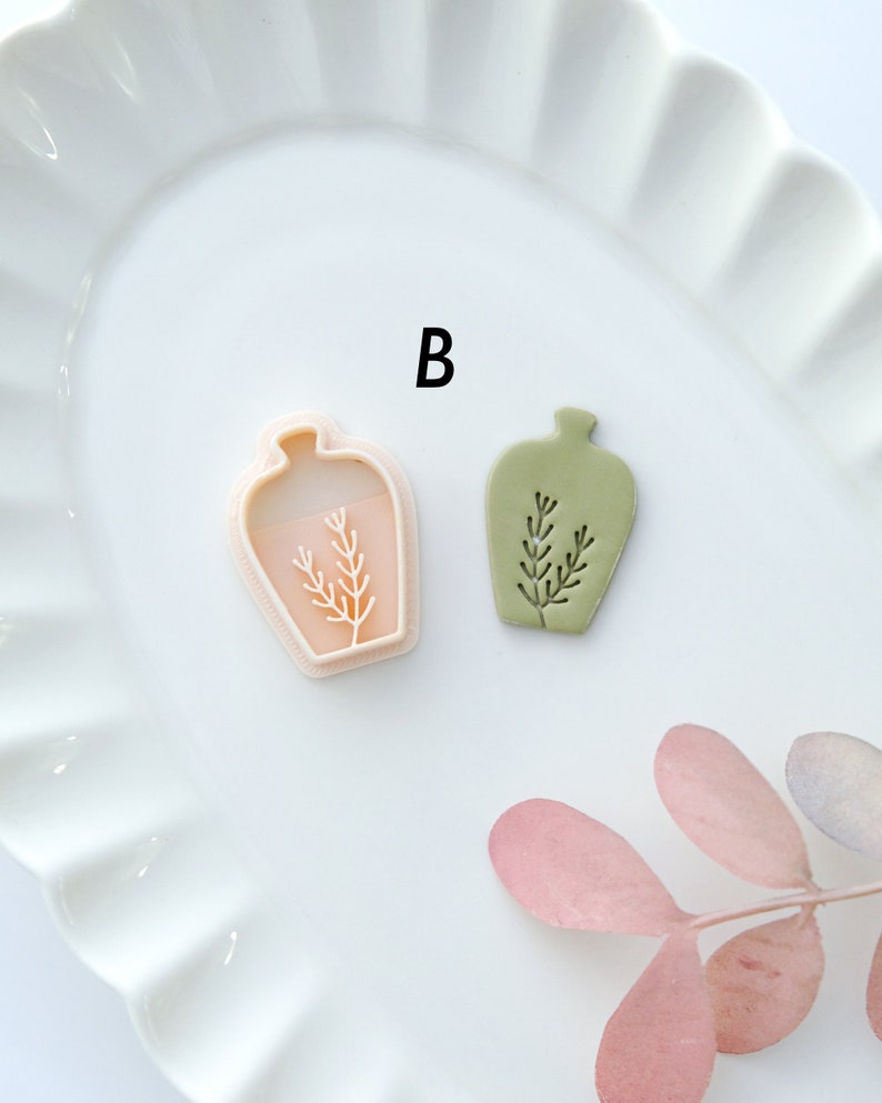 May include: Two ceramic vases with a plant design. The vase on the left is pink and the vase on the right is green. The letter "B" is visible in the background.