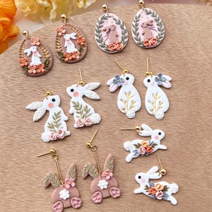 May include: A collection of handmade polymer clay earrings featuring bunny and egg designs. The earrings are in shades of pink, white, and gold with floral accents.
