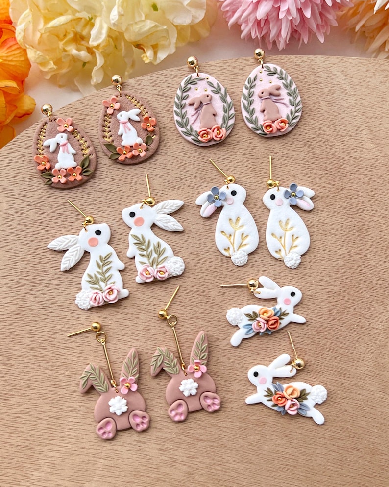May include: A collection of handmade polymer clay earrings featuring bunny and egg designs. The earrings are in shades of pink, white, and gold with floral accents.