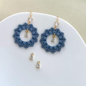 May include: A pair of blue floral earrings with gold-colored hooks and clear, teardrop-shaped charms. The earrings have a circular design with a floral pattern. Two additional charms are also visible.