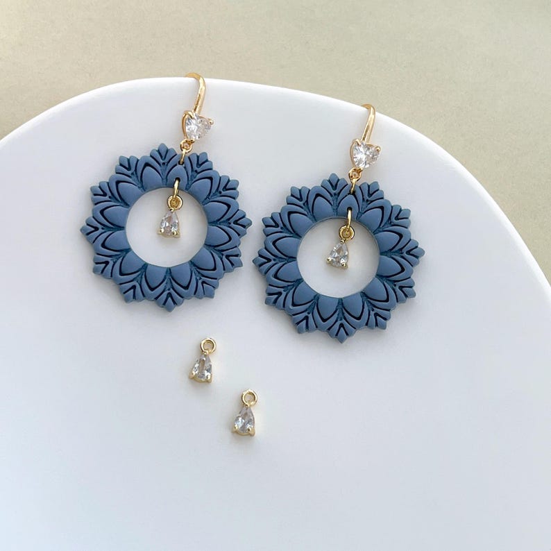 May include: A pair of blue floral earrings with gold-colored hooks and clear, teardrop-shaped charms. The earrings have a circular design with a floral pattern. Two additional charms are also visible.