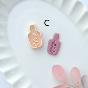 May include: Two cookie cutters in the shape of a bottle with a plant inside. The cutter on the left is white and the cutter on the right is a light purple color. The cutters are on a white plate with a scalloped edge. The letter "C" is written in black on the plate.