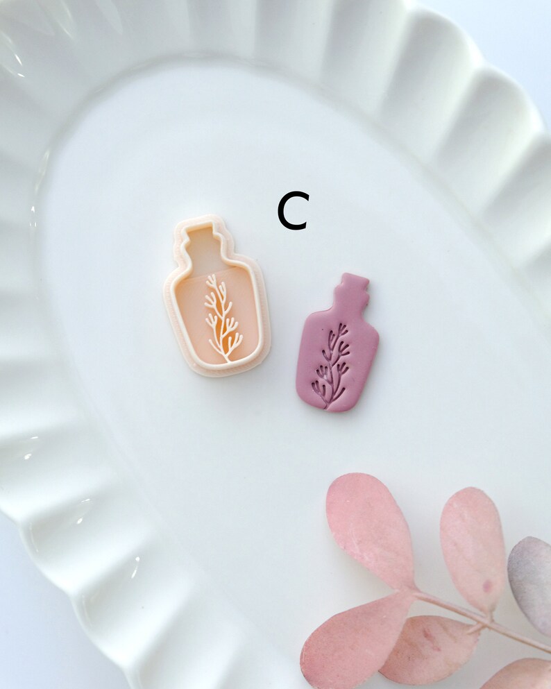 May include: Two cookie cutters in the shape of a bottle with a plant inside. The cutter on the left is white and the cutter on the right is a light purple color. The cutters are on a white plate with a scalloped edge. The letter "C" is written in black on the plate.