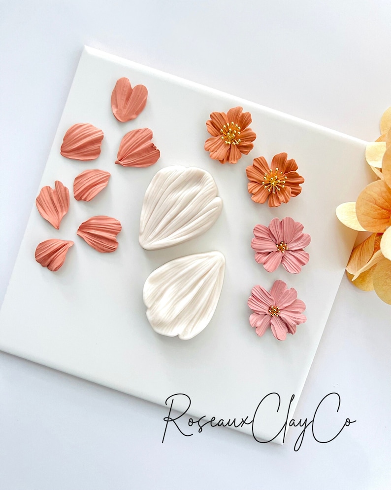 May include: A set of polymer clay flower molds and pre-made flower petals in shades of pink and orange. The molds are white and feature a detailed petal design. The petals are arranged on a white surface with the text "Roseaux Clay Co."