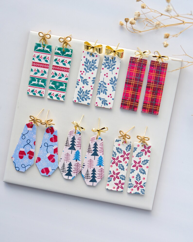 May include: Assortment of eight pairs of Christmas-themed earrings. The earrings are made of metal and feature various holiday patterns, including snowflakes, mittens, Christmas trees, and poinsettias. Each pair has a gold bow charm.