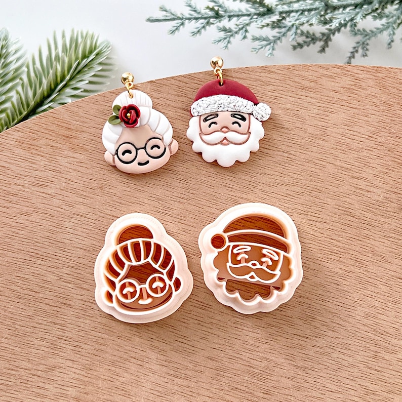 May include: Four Christmas-themed polymer clay earrings, two are finished and two are unfinished. The finished earrings are a Santa Claus and a woman with glasses and a red flower. The unfinished earrings are a Santa Claus and a woman with glasses.
