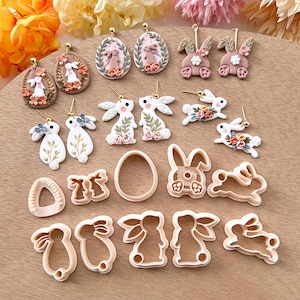 May include: A set of 14 polymer clay bunny and egg earrings and cookie cutters. The earrings are in shades of pink, white, and gold. The cookie cutters are in white and are shaped like bunnies and eggs.